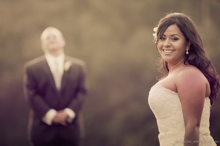 A bride smiles at the camera in the foreground, wearing a white strapless dress. In the blurred background, a man in a suit stands with his hands clasped. The scene is warmly lit, creating a soft, romantic atmosphere.