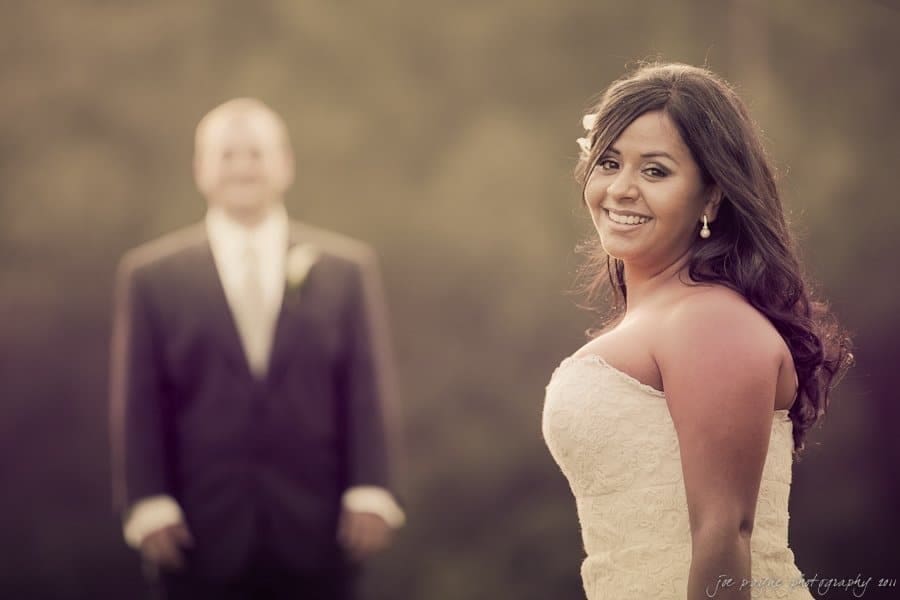 A bride in a white dress smiles at the camera with a blurred groom wearing a suit and tie in the background. The setting is outdoors with a soft focus on the background.