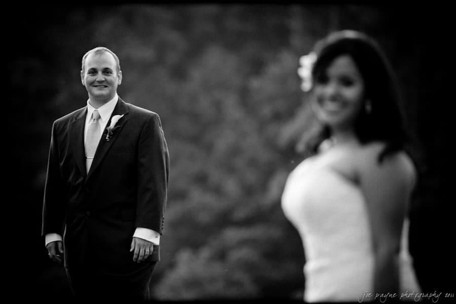 A black-and-white photo of a couple. A man in a suit stands in the background, smiling. In the foreground, a woman in a white dress is out of focus, looking towards the man.