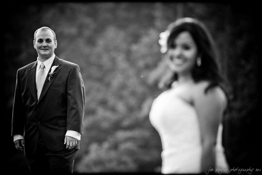 A black and white photo of a bride and groom outdoors. The groom, in a suit with a boutonniere, stands in focus on the left, while the bride, wearing a strapless dress, is blurred in the foreground on the right, smiling.