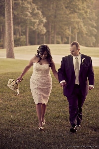 A couple in formal attire holds hands while walking on a grassy area. The woman wears a white lace dress and carries flowers, and the man is in a dark suit with a tie. Trees and a road are visible in the background.
