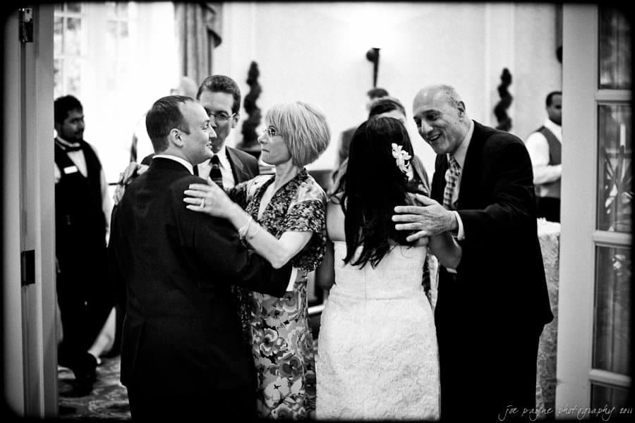 A black and white photo captures five people in a joyous embrace at a social gathering. A bride in a white gown is among them. Two people on the left hold hands. The background shows additional attendees and ornate curtains.