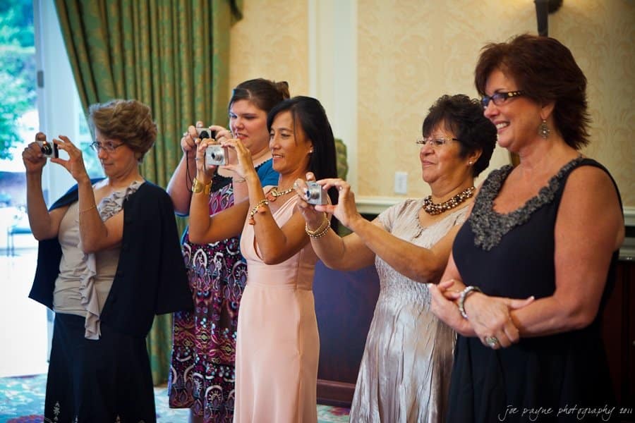 Five women standing in a line, smiling and holding cameras, appearing to take photos. They are dressed in semi-formal attire, with varying hairstyles and dresses. The background features drapes and patterned wallpaper.