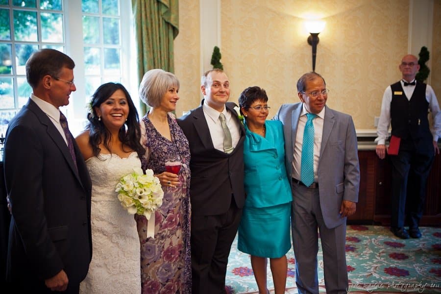 A bride in a white dress holds a bouquet and stands with five people dressed in formal attire. They are smiling and posing together in an elegantly decorated room. A waiter stands in the background.