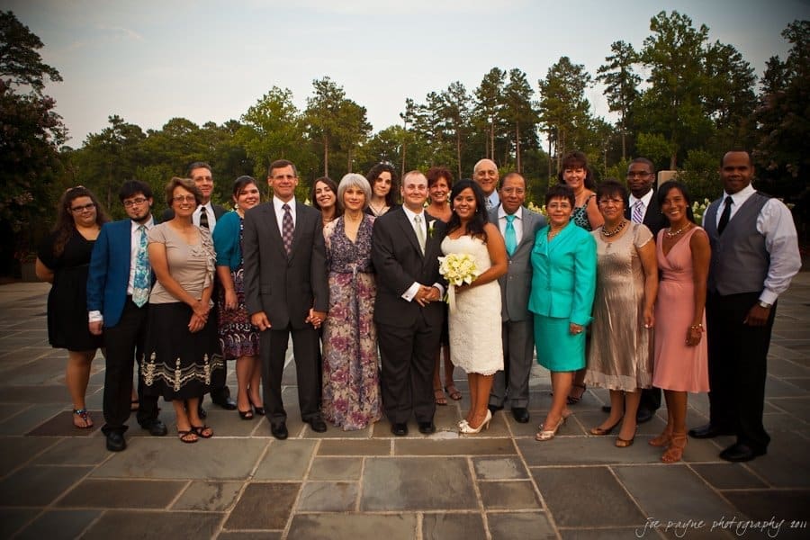 A wedding group photo with a bride in a white dress holding a bouquet and a groom in a dark suit at the center. They are surrounded by smiling family and friends in formal attire, standing on a stone patio with trees in the background.