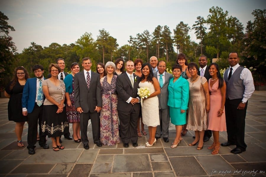 A group photo of people at an outdoor event. The group includes a bride in a white dress holding a bouquet, a groom in a suit, and several others dressed in formal attire. They stand on a stone patio with trees in the background.