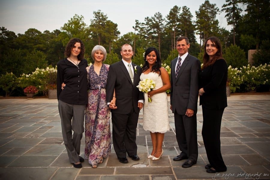 A group of six people, including a bride in a white dress holding flowers and a groom in a suit, stand together on a stone patio. Three women and two men are smiling, surrounded by trees and greenery.