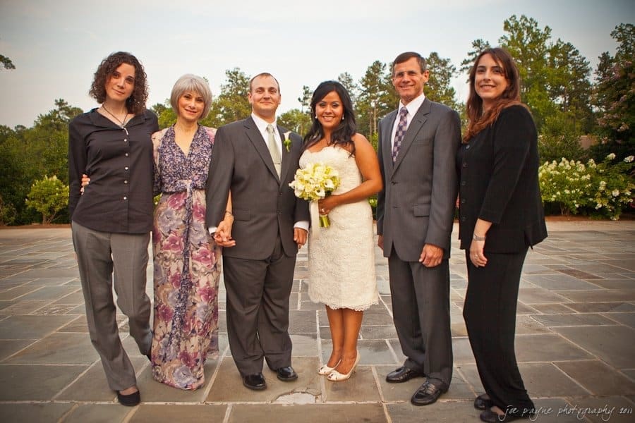 A group photo of six people dressed formally, including the bride in a white dress holding a bouquet and the groom in a suit. They are standing outdoors on a stone patio with greenery in the background.