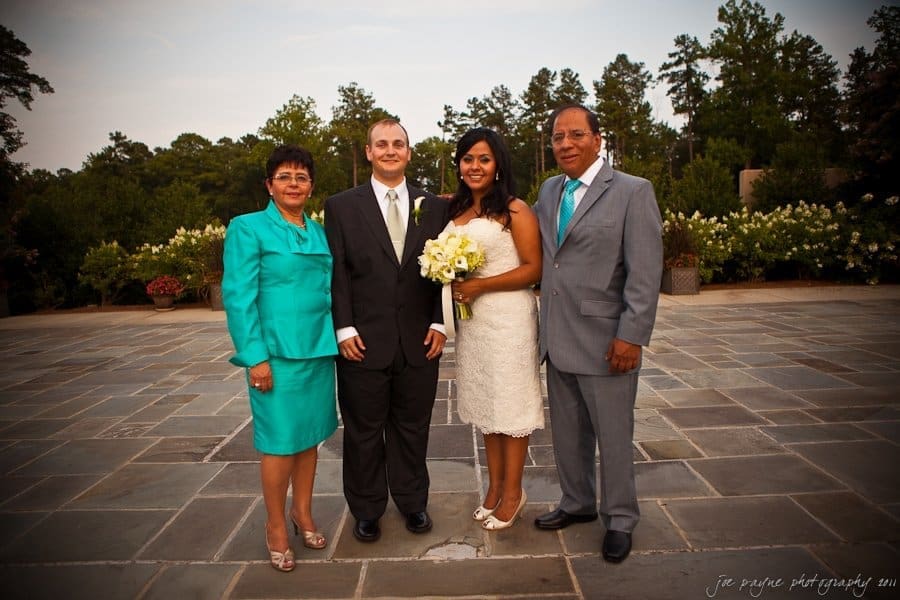 A couple in formal attire stands outdoors on a stone patio with a man and woman. The bride holds a bouquet of white flowers. Trees and potted plants are visible in the background.