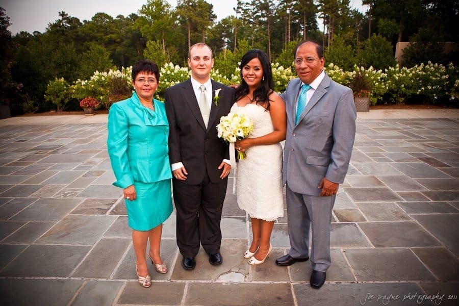 A wedding photo of a bride in a white dress holding a bouquet, standing next to the groom in a black suit. They are flanked by a man in a light blue suit and a woman in a turquoise outfit. Theyre outdoors on a stone pavement, with trees in the background.