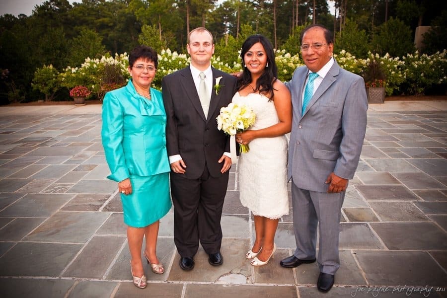 Four people are standing outdoors on a stone patio. A couple in wedding attire is in the center, flanked by two older adults in formal attire. The bride holds a bouquet of yellow and white flowers. Lush greenery is in the background.