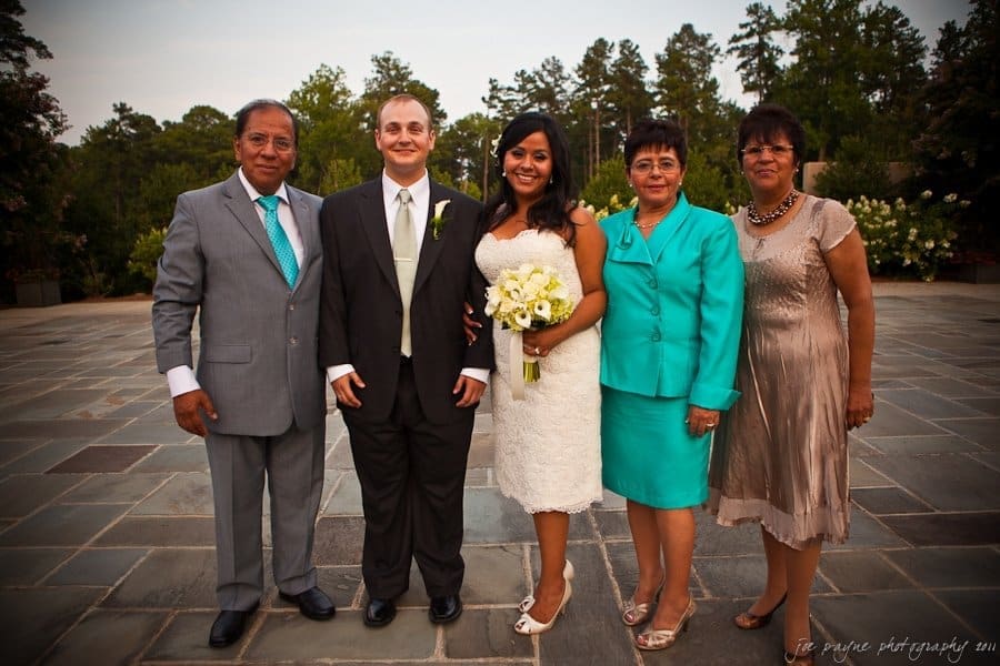 A wedding couple stands with three family members outdoors. The bride wears a white dress and holds a bouquet. The groom is in a suit and tie. Two women wear teal outfits, and one wears a taupe dress. Trees and a tiled patio are in the background.