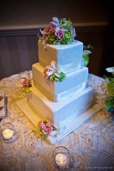 A three-tier square wedding cake adorned with pastel flowers and greenery is set on a textured tablecloth. Two small votive candles glow nearby, adding to the elegant display.