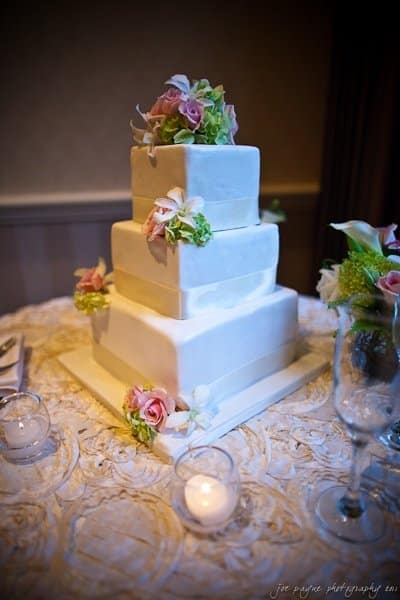 A three-tier square wedding cake with smooth white icing is adorned with pink roses, white lilies, and green hydrangeas. The cake sits on a round, pattern-textured tablecloth with small candles nearby.