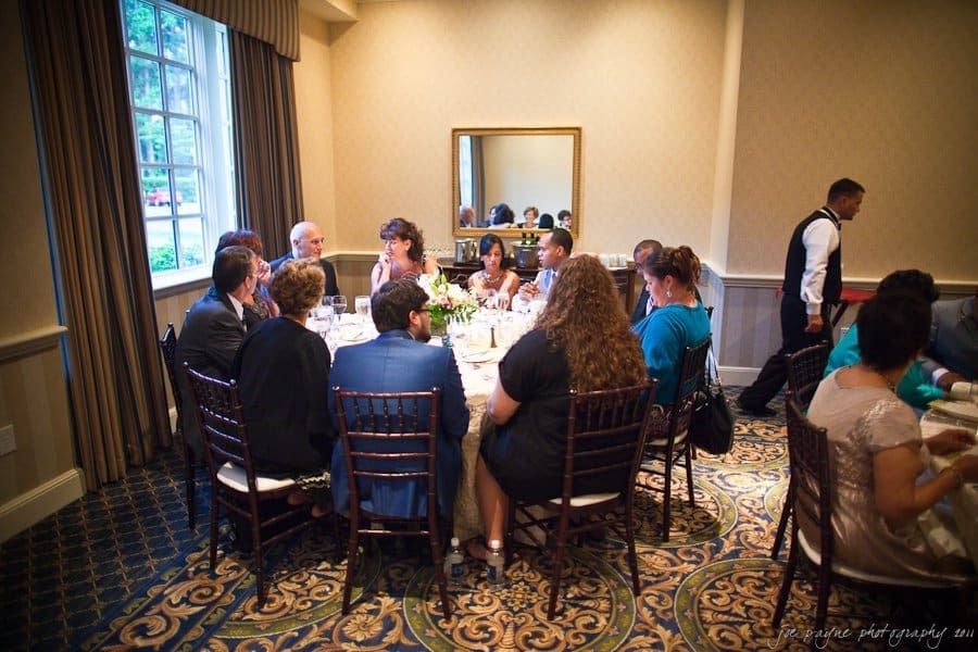 A group of people seated around a dining table in a restaurant or event space. The room is warmly lit, with a large window on the left and a mirror on the wall reflecting part of the scene. A waiter stands nearby attending to the guests.