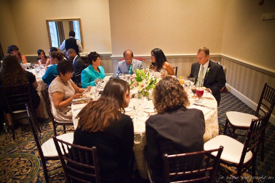 A group of people sit around a round table set for a meal in a warmly lit room. The table is decorated with a floral centerpiece. Everyone is engaged and conversing with each other.