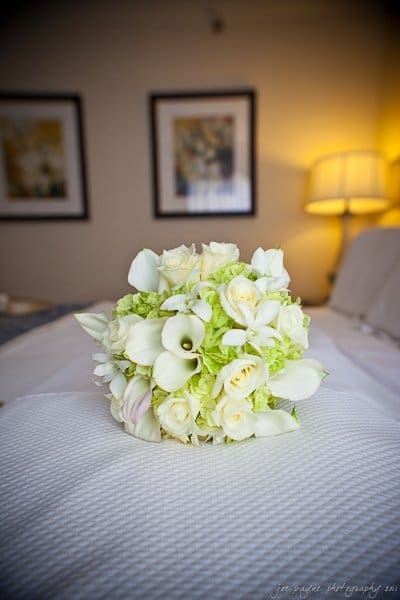A bouquet of white roses, calla lilies, and green hydrangeas sits on a bed with a white textured cover. Two framed pictures and a lit lamp are in the softly blurred background.