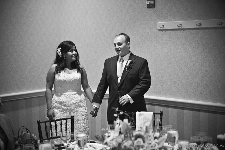 A black and white photo of a couple holding hands and smiling, wearing wedding attire. They are standing near a table decorated with flowers and glassware in a reception setting.