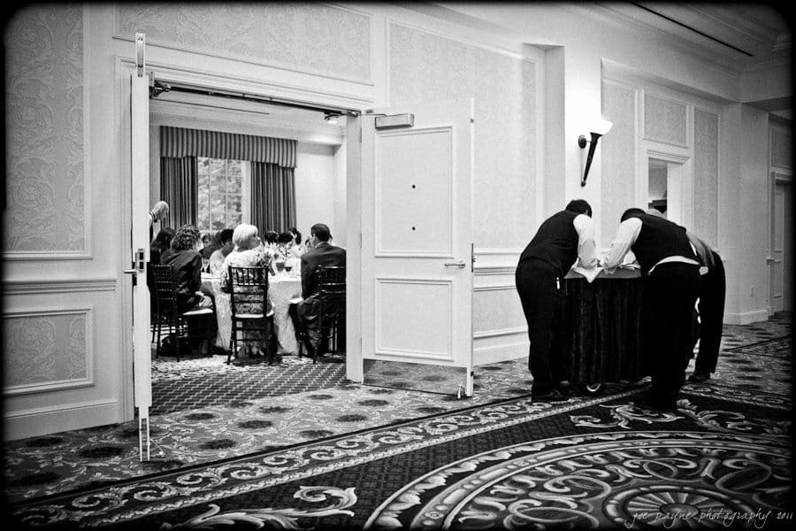 A black and white photo shows a formal event with guests seated at a table through an open doorway. Two staff members in uniforms lean over a table on the right side of the image, possibly preparing or organizing items.