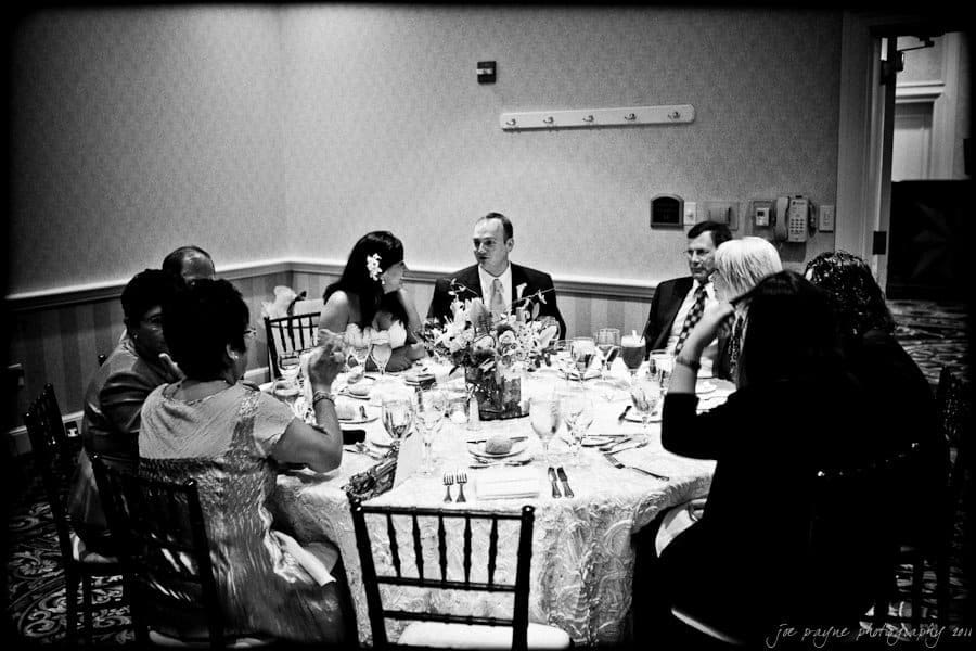 A group of people seated around a round table at a formal event. The table is elegantly set with glasses and a flower centerpiece. The room has a patterned carpet and soft lighting, creating an intimate atmosphere.