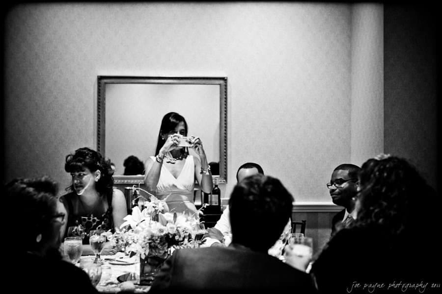 A woman stands at a table taking a photo with a camera, surrounded by seated people engaged in conversation. A mirror hangs on the wall behind them. The scene is in black and white, capturing a lively, social atmosphere.