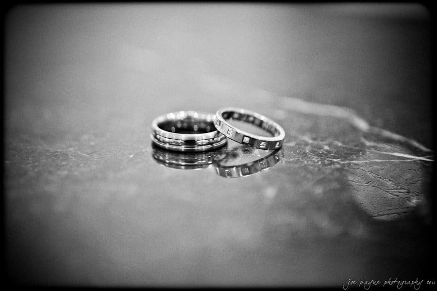 Two wedding rings with intricate designs placed closely on a reflective marble surface, creating a subtle reflection. The image is in black and white, giving it a classic, timeless feel.