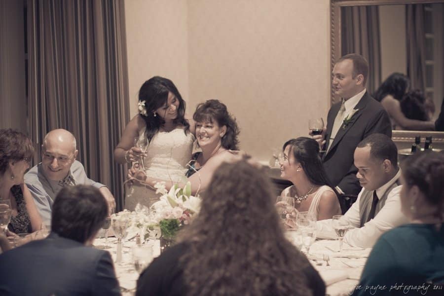 A group of people seated around a dining table, smiling and toasting. Two women are standing, embracing each other joyfully. The room is warmly lit, decorated with flowers, and mirrors adorn the walls.