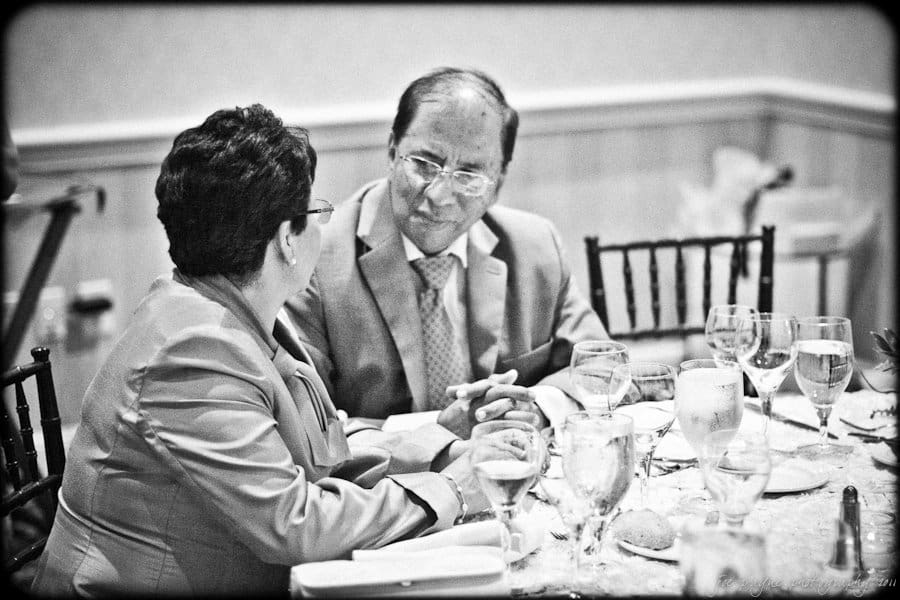 A black-and-white photo of two people seated at a table engaged in conversation. They are dressed formally, with a man in a suit and a woman in a jacket. The table is set with glassware and tableware.