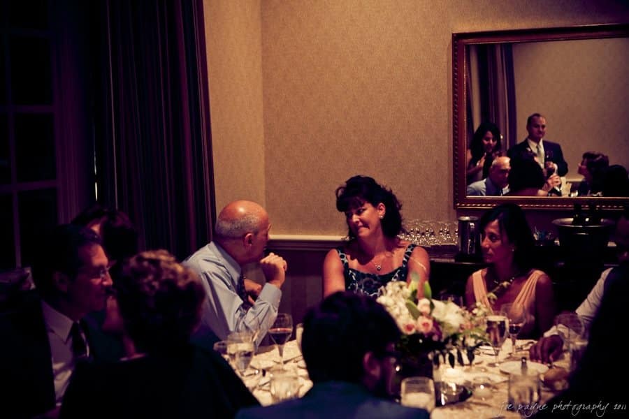 A group of people are seated around a round table at a formal gathering. They are engaged in conversation. Theres a mirror on the wall reflecting additional guests. The table is adorned with a flower arrangement and glasses.