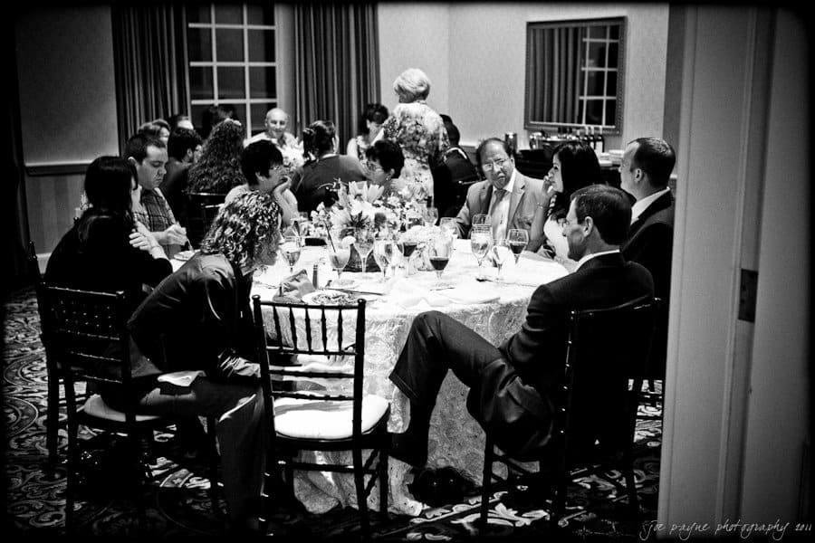 A group of people sit around a large table in a formal dining setting. The table is elegantly set with glassware, flowers, and plates. The room has dark curtains and patterned carpet, creating an intimate atmosphere.