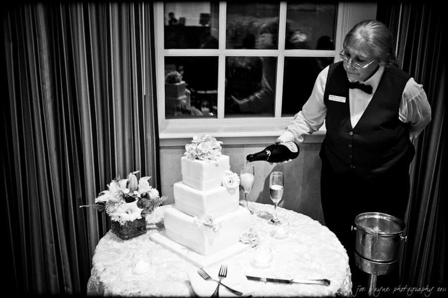 A server in formal attire pours champagne next to a three-tiered wedding cake adorned with flowers. The table is elegantly set with a vase of flowers, a knife, and champagne glasses.