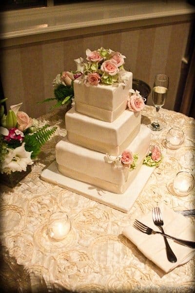 A three-tiered square wedding cake with white icing and pink roses on top. The cake sits on a decorated table with candles, a bouquet, a folded napkin, utensils, and a glass of champagne.
