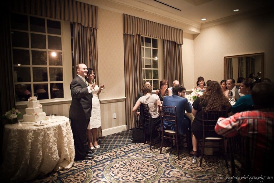 A bride and groom stand near a table with a wedding cake, raising glasses in a toast. They face seated guests in a warmly lit reception room with patterned carpeting and draped windows.