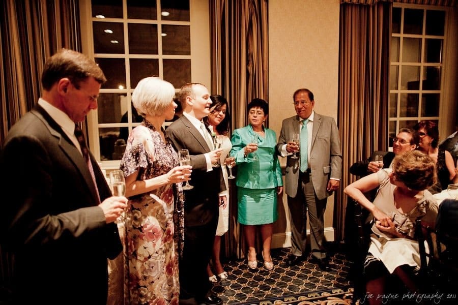 A group of people dressed in formal attire gather indoors, holding champagne glasses. They appear to be celebrating, with some smiling and engaged in conversation. The setting has elegant decor with large windows and curtains.