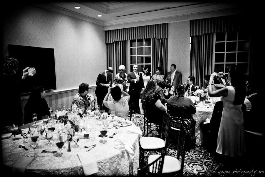 A black and white photo of a formal indoor gathering. Guests sit at decorated tables with wine glasses, while others stand near the windows. Some people are taking photos, and a few are giving a speech or presentation in front of the group.