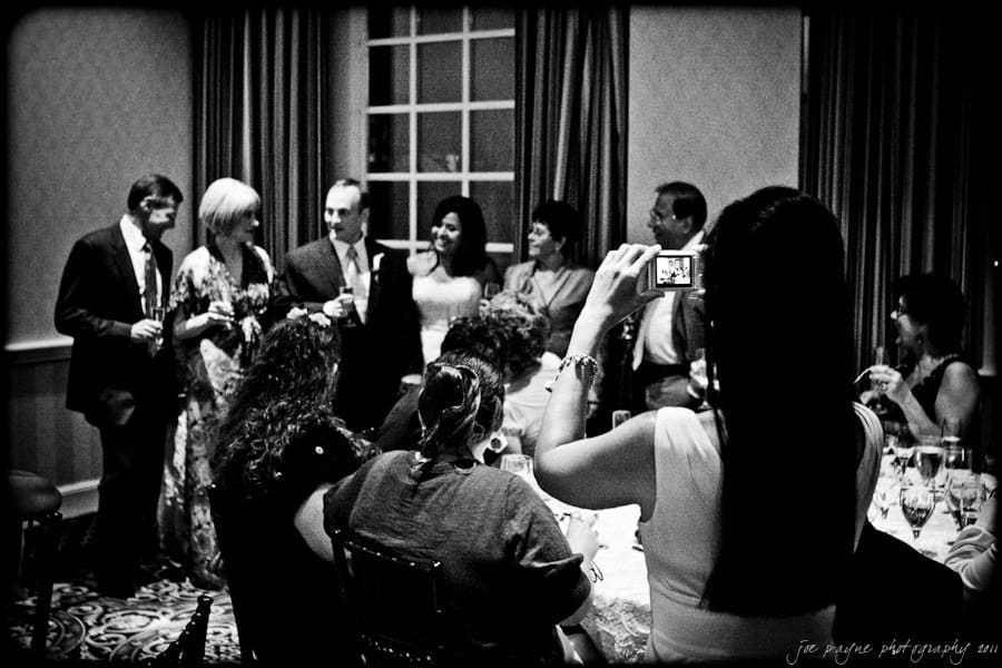 A black-and-white photo of a group of six people standing and posing for a picture in a room. An individual in the foreground is taking the picture with a camera. Seated guests are partially visible at a table. The setting appears formal.