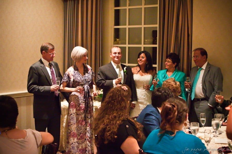 A group of six people in formal attire stands together in a warmly lit room, holding glasses as if giving a toast. There are elegantly set tables and guests seated in the foreground. The window behind them suggests an evening event.