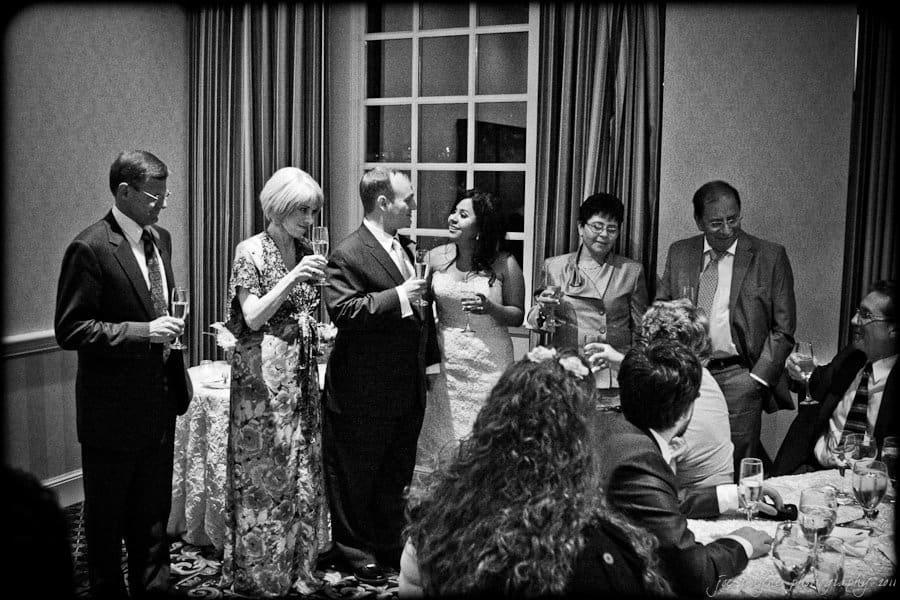 A black and white photo of a wedding reception shows a bride and groom holding champagne glasses. They stand among guests, who are also holding drinks, engaged in conversation and celebration. The setting is an elegant room with a large window.