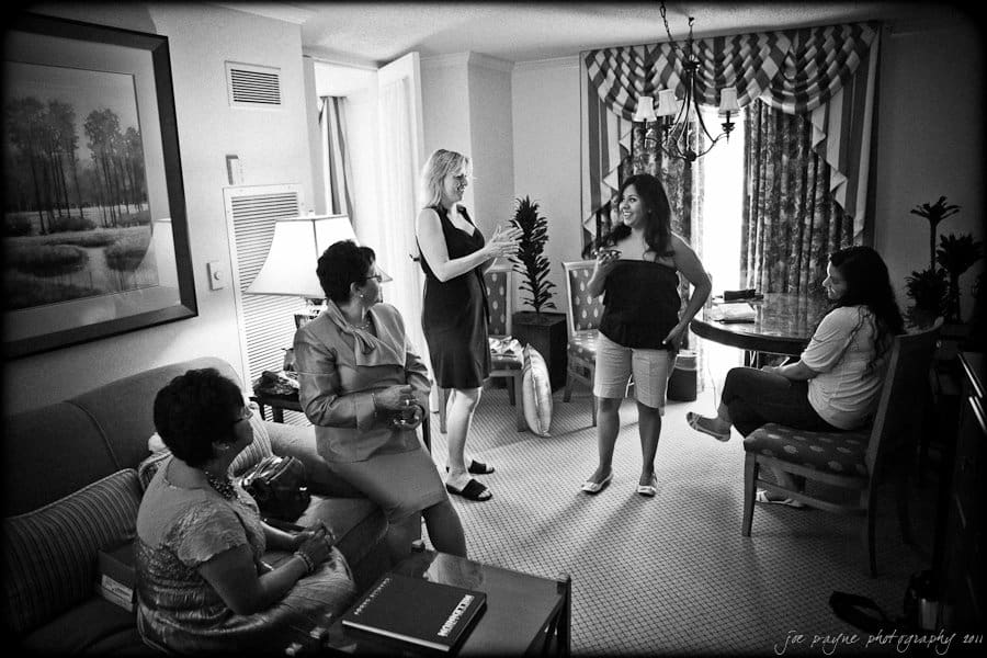 A black and white photo of five women in a living room. Two women are standing and conversing, while the other three are seated, engaged in conversation. The room is decorated with a painting, curtains, and a table with a laptop.