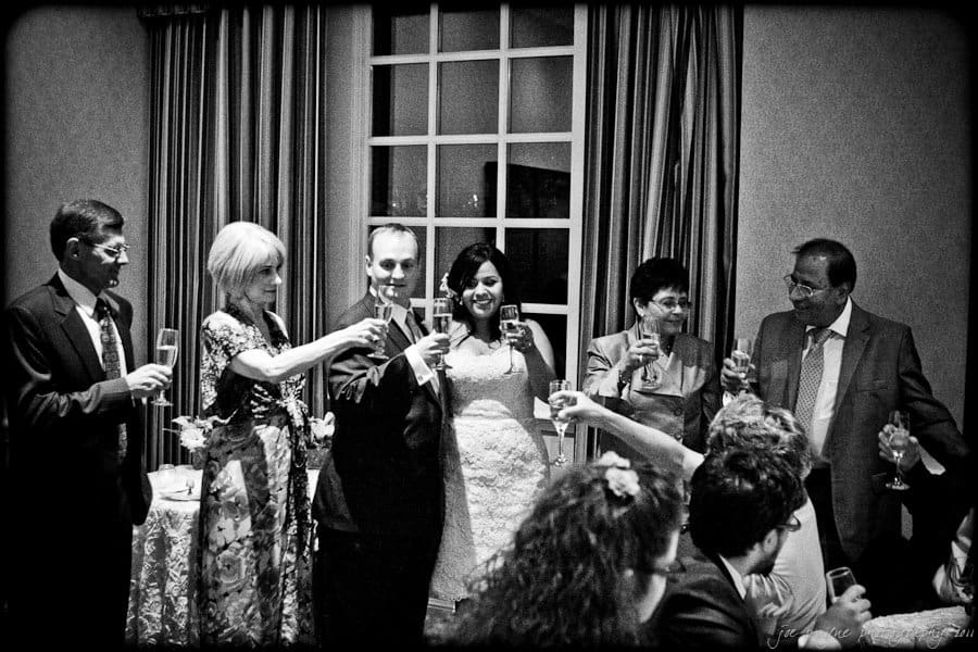 A group of people stand together inside a room holding glasses in a toast. They are smiling and dressed in formal attire. A window with curtains is in the background, and other people are seated in the foreground. The image is in black and white.