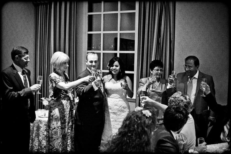 A black-and-white photo of a wedding reception. A bride and groom are surrounded by family and friends, raising champagne glasses for a toast. They stand in front of a window with curtains, with a table of gifts visible nearby.