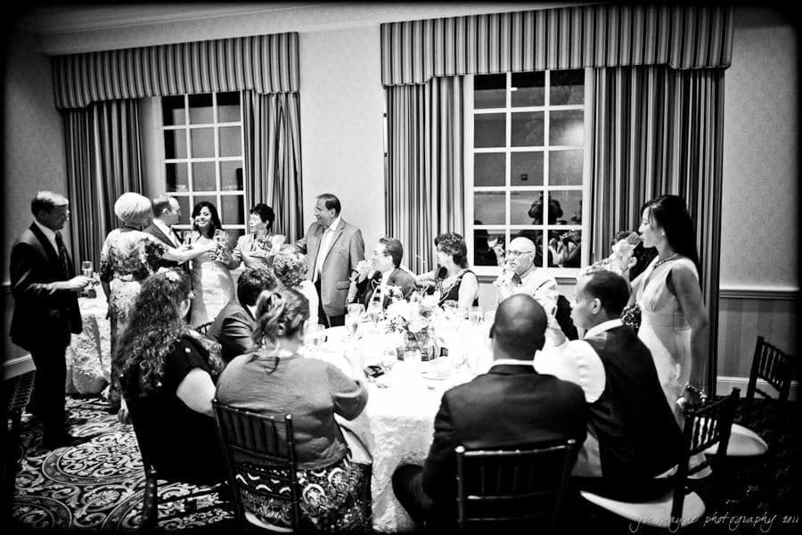 A black and white photo of a wedding reception. Guests are seated around a table with floral centerpieces. The bride and groom are standing, toasting with glasses. Large windows and decorative curtains are in the background.