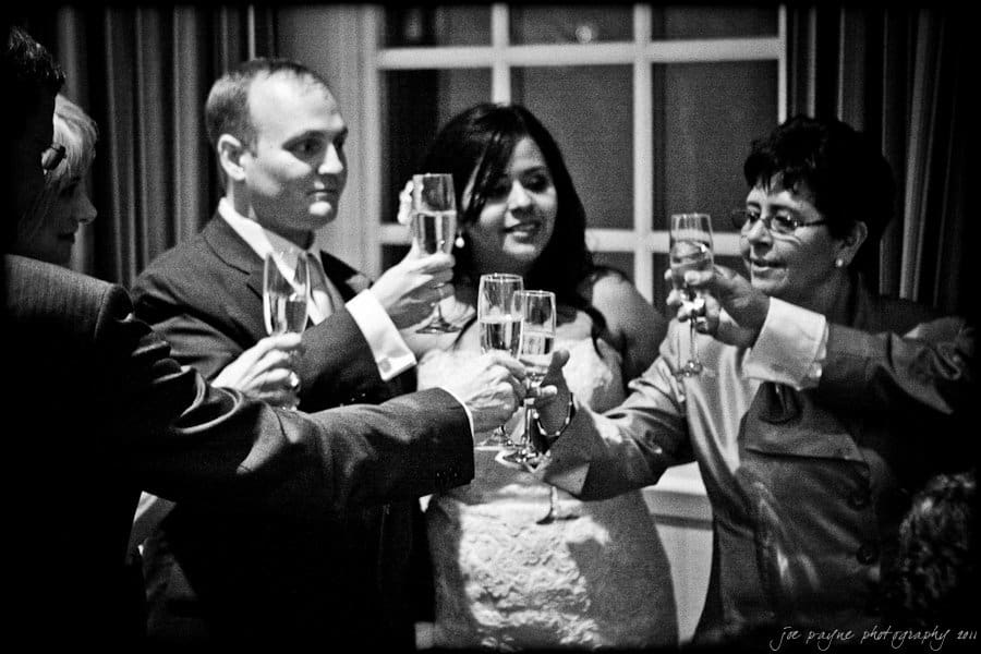 A group of people in formal attire raise champagne glasses in a toast. They are gathered in a dimly lit room with a window in the background. The mood appears celebratory and joyful.