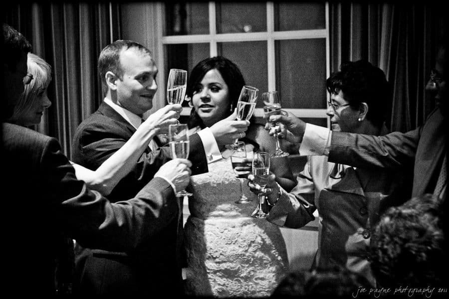 A group of people dressed in formal attire raise their glasses in a celebratory toast in a dimly lit room by a window.