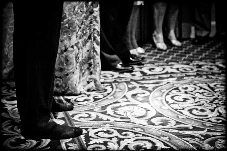 A black and white image showing a group of people standing on an ornate patterned carpet. Visible are their legs and feet, with some wearing formal shoes and dresses. The scene suggests a formal event or gathering.