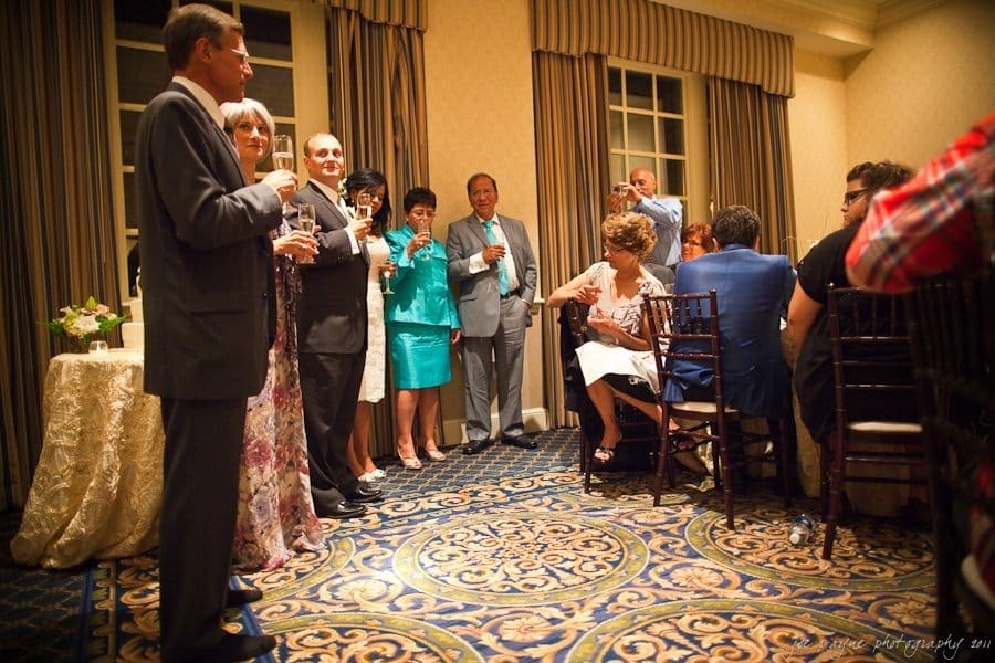 A group of people dressed in formal attire are gathered in a decorated room, holding drinks. Some are standing and others are seated at a table. The setting suggests a social event or celebration, with patterned carpet and draped windows.