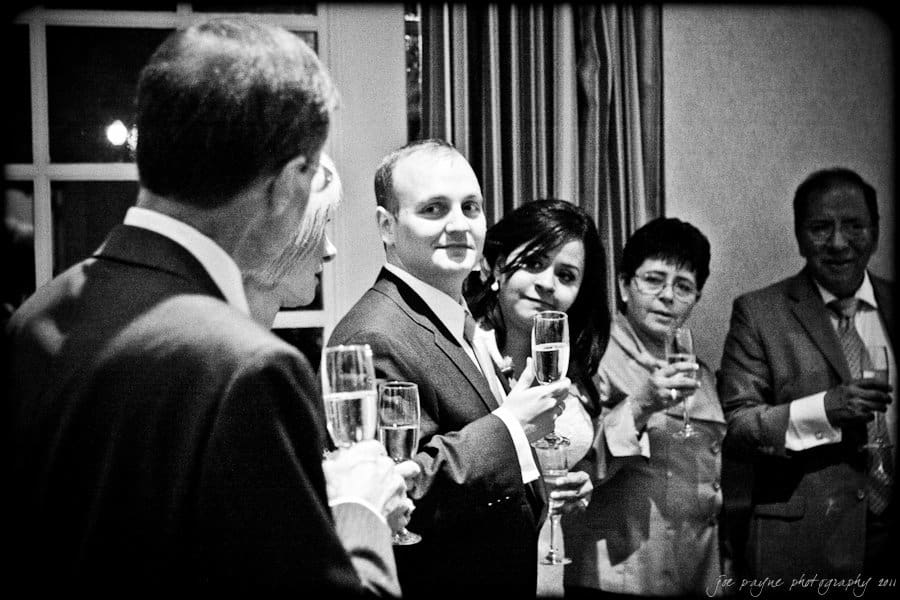 A black and white photo of a group of people holding champagne glasses, standing in an elegant room. They are dressed formally and appear to be toasting or celebrating an occasion.