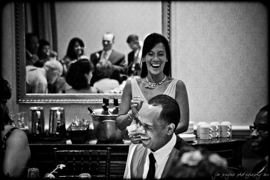 A black-and-white photo of a lively dinner party. A man is laughing, while a woman stands nearby, laughing with him. Several people are gathered in the background, smiling and enjoying the atmosphere.