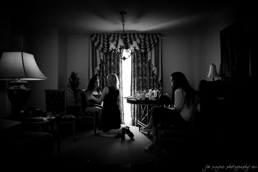 A black and white image of a dimly lit room with three women. One sits on a chair, another kneels in front of her, and the third is seated at a table with makeup products. Drapes cover the window, and a chandelier hangs overhead.