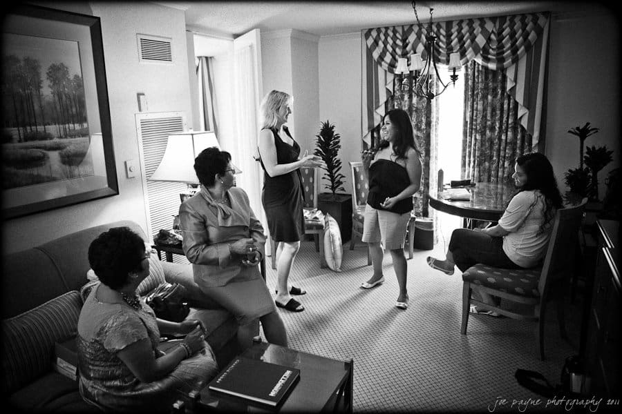 A group of women are gathered in a living room, engaged in conversation. Two women stand in the center, talking, while three others sit and listen. The room is decorated with a painting, curtains, and a lamp.